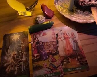 May include: Two lit candles, a crystal ball, a green crystal, a red stone, a white candle in a seashell, a bundle of sage, and two tarot cards, one showing the Ten of Pentacles and the other showing the Three of Chalices.