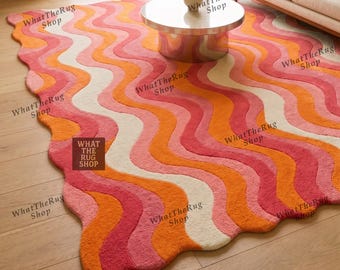 May include: A large area rug featuring a wavy, multi-coloured pattern in shades of orange, pink, red, and cream. The rug has an irregular, organic shape with curved edges and a textured surface, displaying the text WHAT THE RUG SHOP on a label.