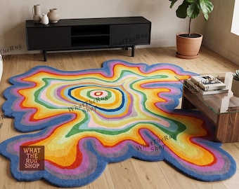 Puede incluir: Una alfombra de forma orgánica irregular con un patrón concéntrico multicolor vibrante de bandas onduladas en azul, verde, amarillo, naranja y rosa. La alfombra muestra un diseño circular central que se irradia hacia afuera en formas fluidas.