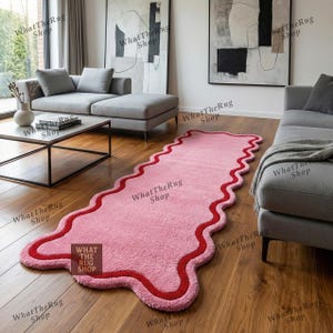 Puede incluir: Una alfombra rosa con forma ondulada e irregular y borde rojo. La alfombra está colocada sobre un suelo de madera en un salón. El texto "What The Rug Shop" es visible en la imagen.