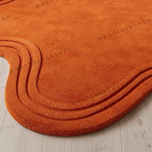 May include: Close-up of an orange, wavy-edged rug. The rug has a textured surface and a three-layered, curved design along the edge. The words "WHATTHERUGSHOP" are faintly visible on the rug's surface.