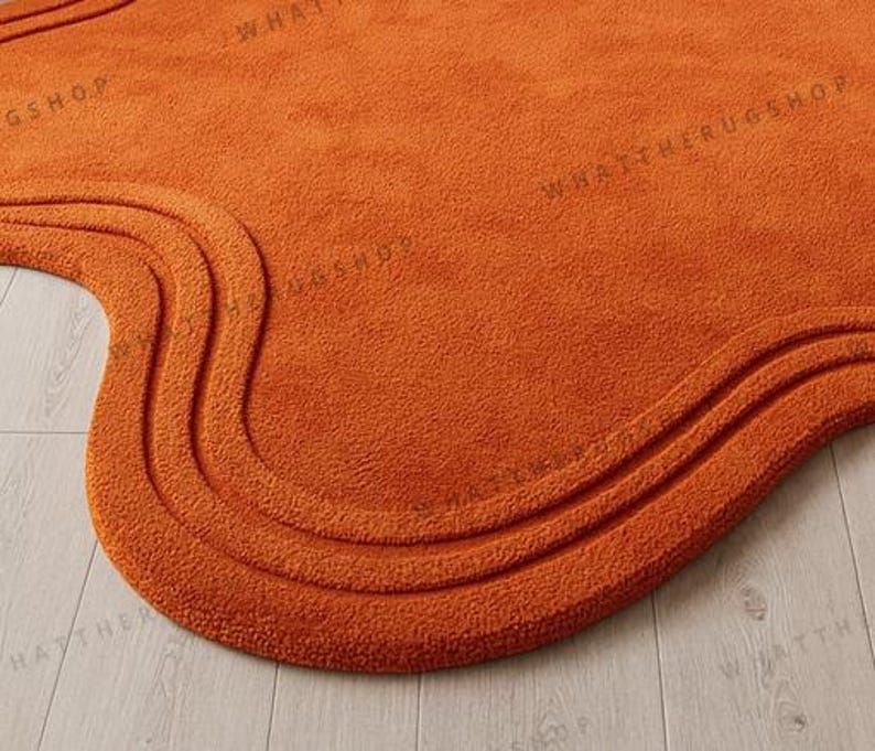 May include: Close-up of an orange, wavy-edged rug. The rug has a textured surface and a three-layered, curved design along the edge. The words "WHATTHERUGSHOP" are faintly visible on the rug's surface.