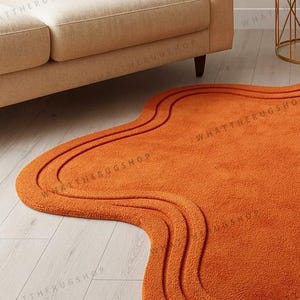 May include: An orange, wavy-shaped rug with a textured surface. The rug has a unique, organic design with three curved lines that follow the shape of the rug. The rug is placed on a light-coloured wooden floor, near a beige sofa.