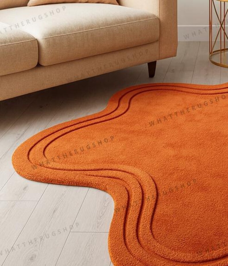 May include: An orange, wavy-shaped rug with a textured surface. The rug has a unique, organic design with three curved lines that follow the shape of the rug. The rug is placed on a light-coloured wooden floor, near a beige sofa.