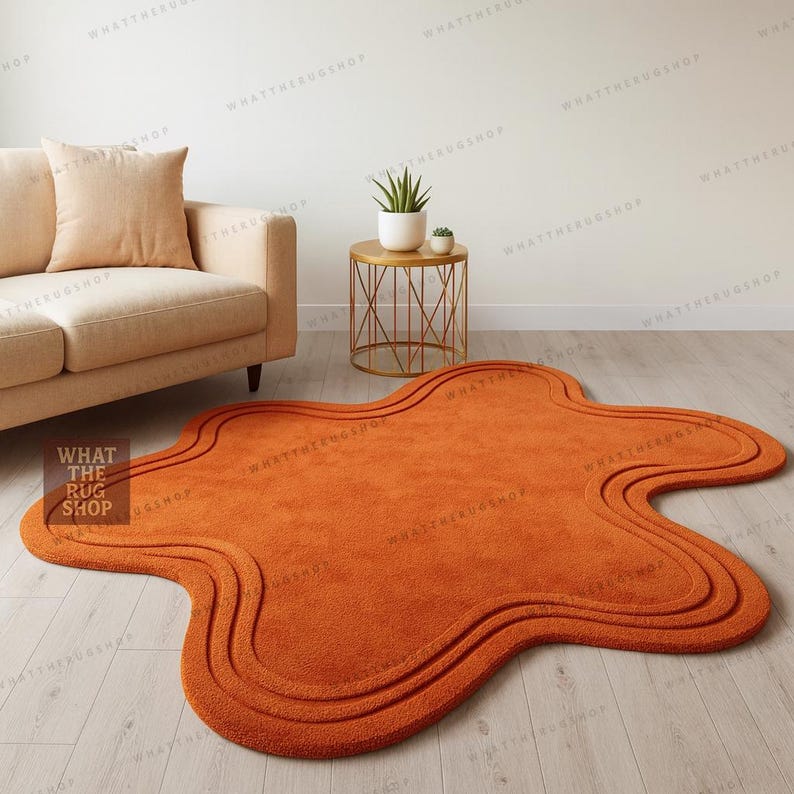 May include: An orange rug with an unusual, flowing shape. The rug has a textured surface and a three-layered border design. It's placed on a light wooden floor. The text "WHAT THE RUG SHOP" is visible on the rug.