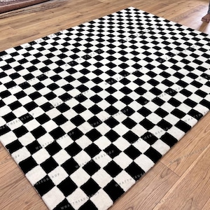 Può includere: Un grande tappeto rettangolare con un motivo a scacchiera bianco e nero. Il tappeto è realizzato in un materiale morbido ed è posizionato su un pavimento in legno. Il tappeto ha il testo "WHAT THE RUG SHOP" stampato.