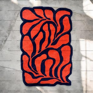 Könnte beinhalten: Ein rechteckiger Teppich mit einem auffälligen Design. Der Teppich hat ein leuchtend orangefarbenes Muster auf einem dunkelblauen Hintergrund. Das Design ist abstrakt, mit fließenden, organischen Formen. Die Worte "WHATTHERUGSHOP" wiederholen sich.