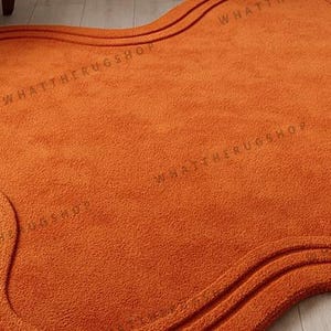 May include: An orange rug with a wavy, organic shape. The rug has a textured surface and a darker orange border with two parallel lines. The text "WHATTHERUGSHOP" is printed on the rug.