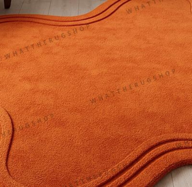 May include: An orange rug with a wavy, organic shape. The rug has a textured surface and a darker orange border with two parallel lines. The text "WHATTHERUGSHOP" is printed on the rug.