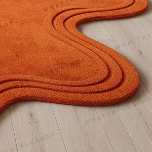 May include: An orange, abstract-shaped rug with a wavy design. The rug has a textured surface and a thick pile. The rug is placed on a light-coloured wooden floor. The rug's shape is reminiscent of a free-form design.