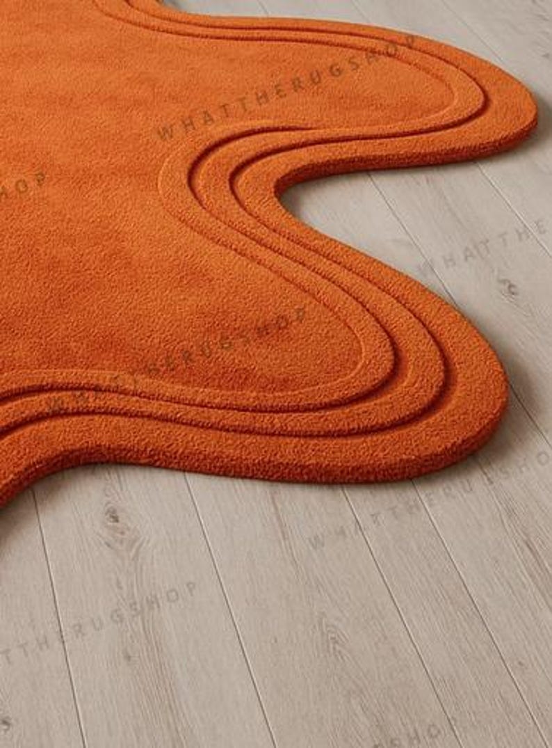 May include: An orange, abstract-shaped rug with a wavy design. The rug has a textured surface and a thick pile. The rug is placed on a light-coloured wooden floor. The rug's shape is reminiscent of a free-form design.