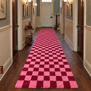 May include: A long, rectangular rug featuring a pink and burgundy checkerboard design. The rug is laid in a hallway with dark wood flooring and white paneling. The rug's pattern provides a striking visual contrast to the hallway's traditional features.