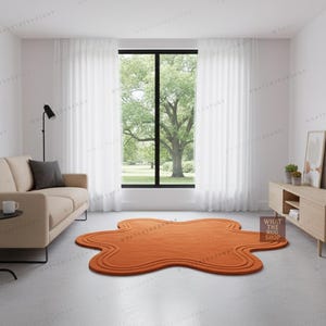 May include: An orange, abstract-shaped rug with a wavy border is placed on a light grey floor. The rug is in a living room setting with a beige sofa, a black lamp, and a large window with white curtains. The room has a modern, minimalist design.