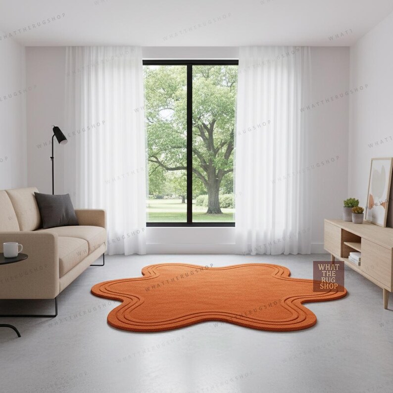 May include: An orange, abstract-shaped rug with a wavy border is placed on a light grey floor. The rug is in a living room setting with a beige sofa, a black lamp, and a large window with white curtains. The room has a modern, minimalist design.
