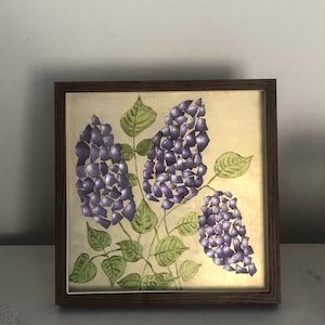 Può includere: Un'opera d'arte incorniciata con un dipinto di fiori viola con foglie verdi su uno sfondo crema.