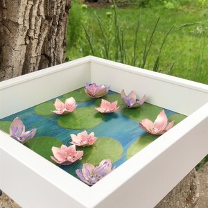 Op de afbeelding: Een wit frame met een blauwe en groene achtergrond met roze en paarse papieren bloemen. De bloemen zijn gerangschikt in een patroon dat lijkt op een vijver met waterlelies.