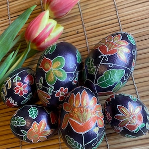 Op de afbeelding: Zes donkerpaarse paaseieren versierd met ingewikkelde bloemmotieven in tinten oranje, goud en groen. De eieren zijn gerangschikt op een geweven bamboemat.