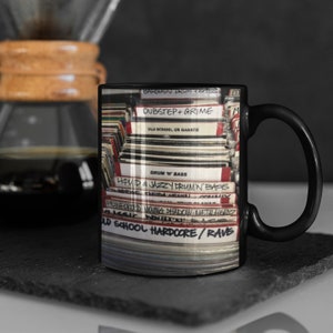 May include: Black coffee mug featuring a stack of vinyl records with handwritten labels. The labels include genres like Dubstep + Grime, Drum 'N' Bass, and Old School Hardcore/Rave. A coffee maker is in the background.