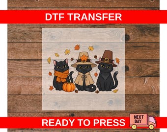 May include: DTF TRANSFER and READY TO PRESS text on a wooden background. The image features three black cats wearing fall-themed attire, including hats, scarves, and a cape. A pumpkin and autumn leaves are also present.