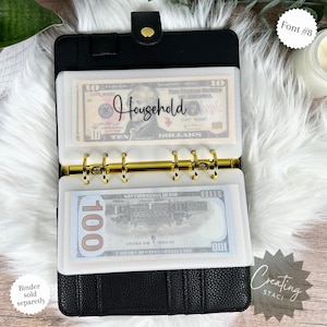 May include: A black leather binder with gold rings and clear plastic pockets for cash. The pockets are labeled "Household" and contain US currency. The binder is closed and has a textured surface.
