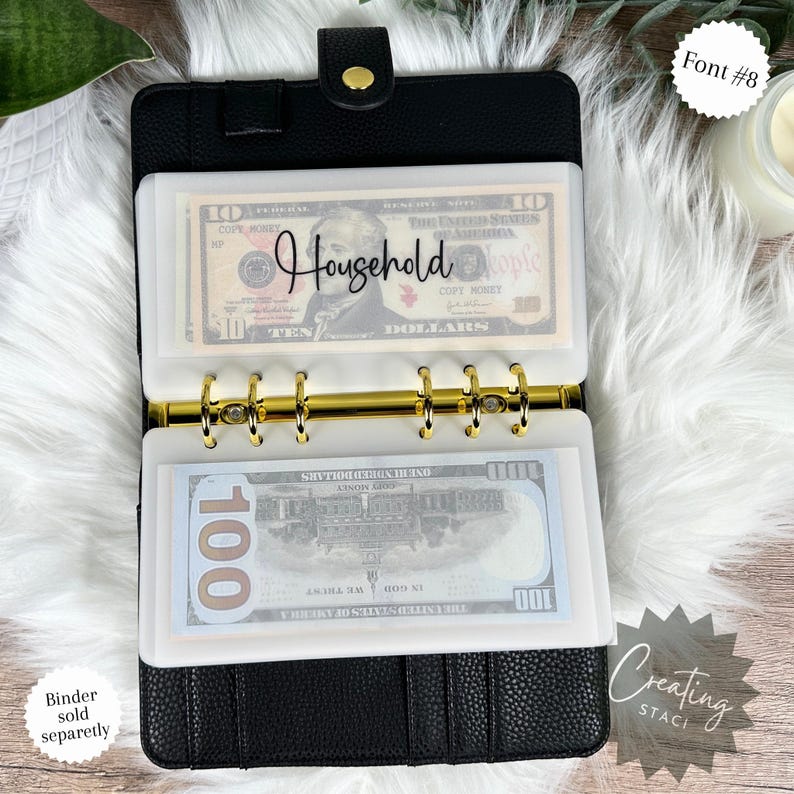 May include: A black leather binder with gold rings and clear plastic pockets for cash. The pockets are labeled "Household" and contain US currency. The binder is closed and has a textured surface.