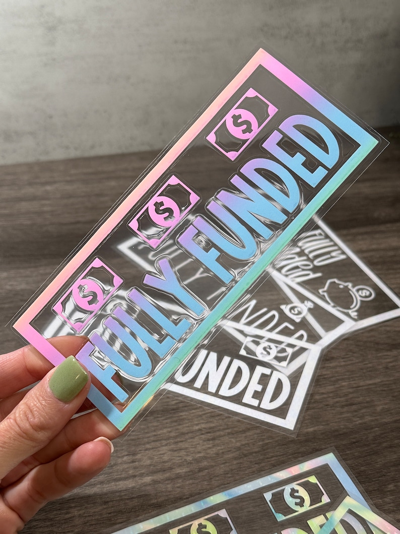 Puede incluir: Una pegatina rectangular con un borde hologr&aacute;fico y el texto "FULLY FUNDED" en letras grandes y negritas. El dise&ntilde;o incluye peque&ntilde;os iconos de billetes de d&oacute;lar con el s&iacute;mbolo de d&oacute;lar. La pegatina tiene un fondo transparente y un acabado reflectante.