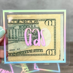 May include: A clear plastic cash envelope featuring a holographic border and the word "gas" in pink script lettering. Inside the envelope is a ten-dollar bill. The design is rectangular with rounded corners and a transparent finish.