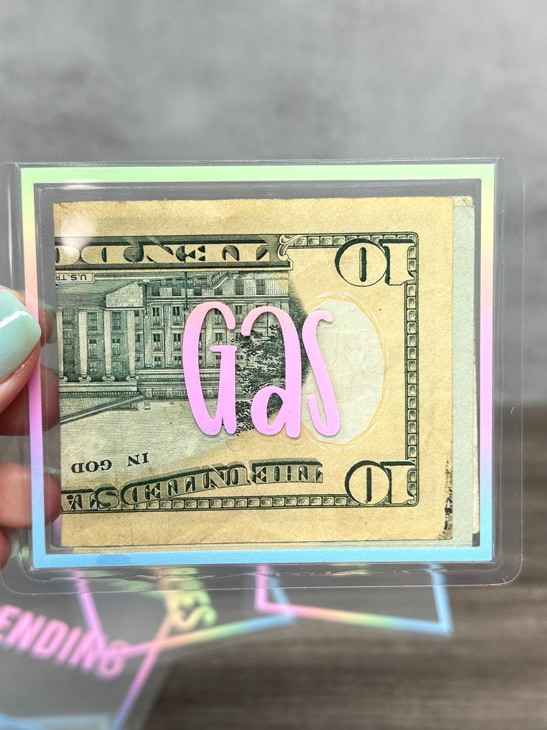 May include: A clear plastic cash envelope featuring a holographic border and the word "gas" in pink script lettering. Inside the envelope is a ten-dollar bill. The design is rectangular with rounded corners and a transparent finish.