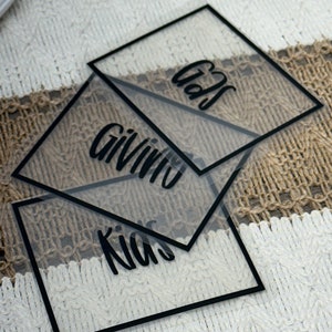 May include: Three square, transparent acrylic signs with black borders and black lettering. The signs feature the words "Gifts," "Giving," and "Kids" in a cursive script font. The clear material allows the underlying surface to be visible.