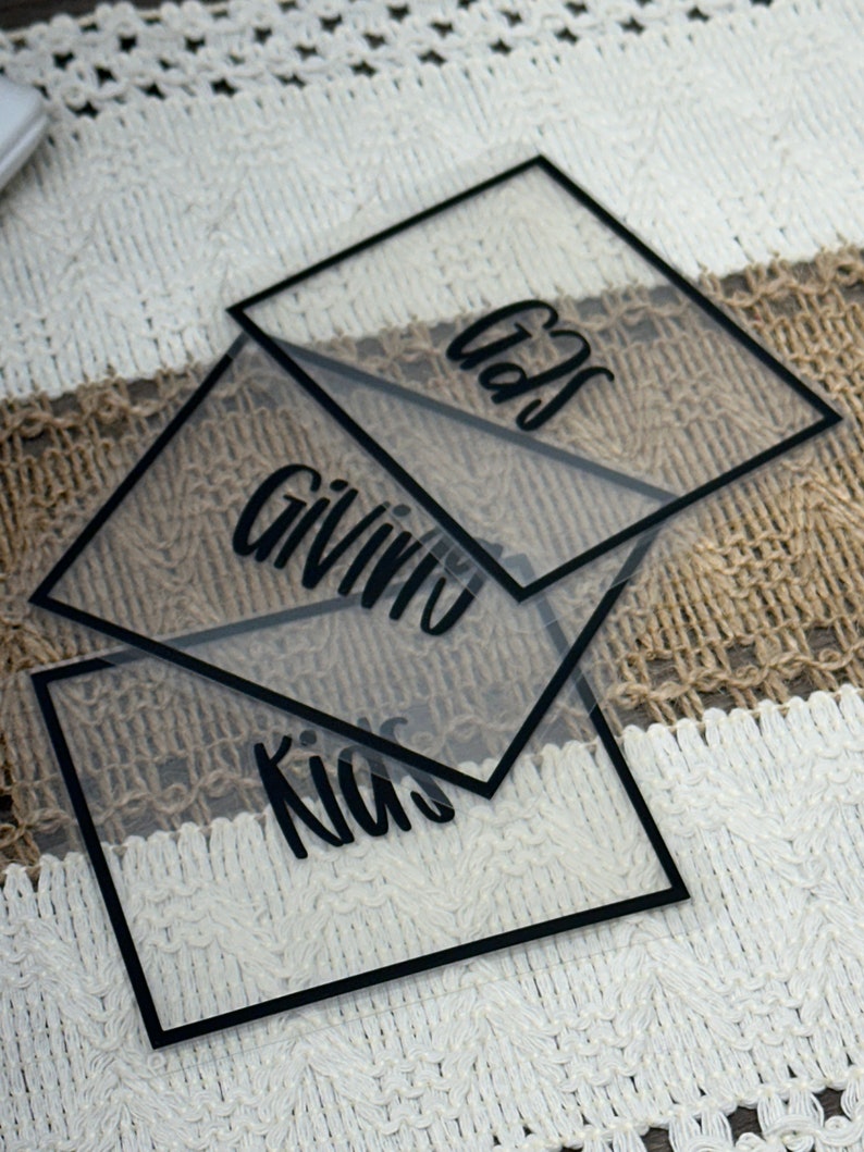 May include: Three square, transparent acrylic signs with black borders and black lettering. The signs feature the words "Gifts," "Giving," and "Kids" in a cursive script font. The clear material allows the underlying surface to be visible.