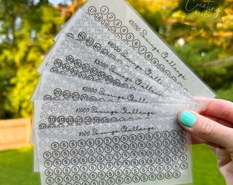 May include: A set of six transparent sheets with a savings challenge printed on each. The sheets are labeled with different dollar amounts, including $500, $1000, $2000, $3000, $4000, and $5050. Each sheet has a grid of circles with numbers inside, representing the amount to save each time.