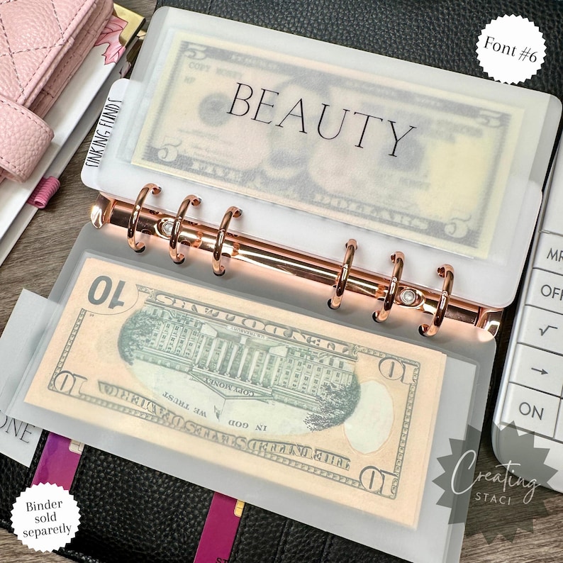 May include: A white plastic binder with a clear plastic pocket that holds cash. The pocket is labeled "BEAUTY" in black text. The binder has a rose gold ring mechanism.
