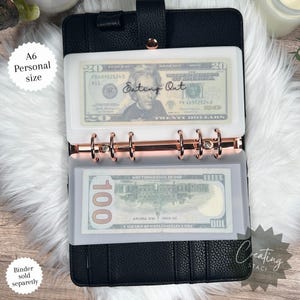 May include: A black A6 personal size binder with a clear plastic pocket for cash. The pocket has the text "Eating Out" written on it. The binder has a rose gold ring mechanism. There are two bills in the pocket, a $20 bill and a $100 bill.