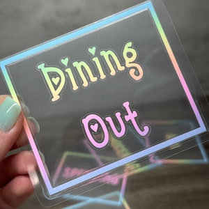 May include: A square, transparent sticker featuring a holographic border and the text "Dining Out" in a gradient font. The letters "i" in "Dining" and "o" in "Out" contain small heart cutouts. The design reflects a rainbow color spectrum.