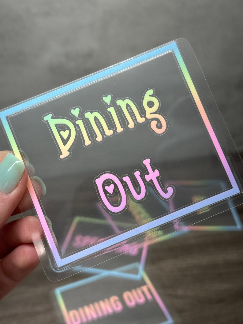 May include: A square, transparent sticker featuring a holographic border and the text "Dining Out" in a gradient font. The letters "i" in "Dining" and "o" in "Out" contain small heart cutouts. The design reflects a rainbow color spectrum.