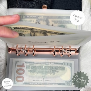 May include: A white plastic cash envelope with a rose gold binder ring system. The envelope has the words "Cash Out" written on it. The envelope is open at the top and has a $100 bill inside.