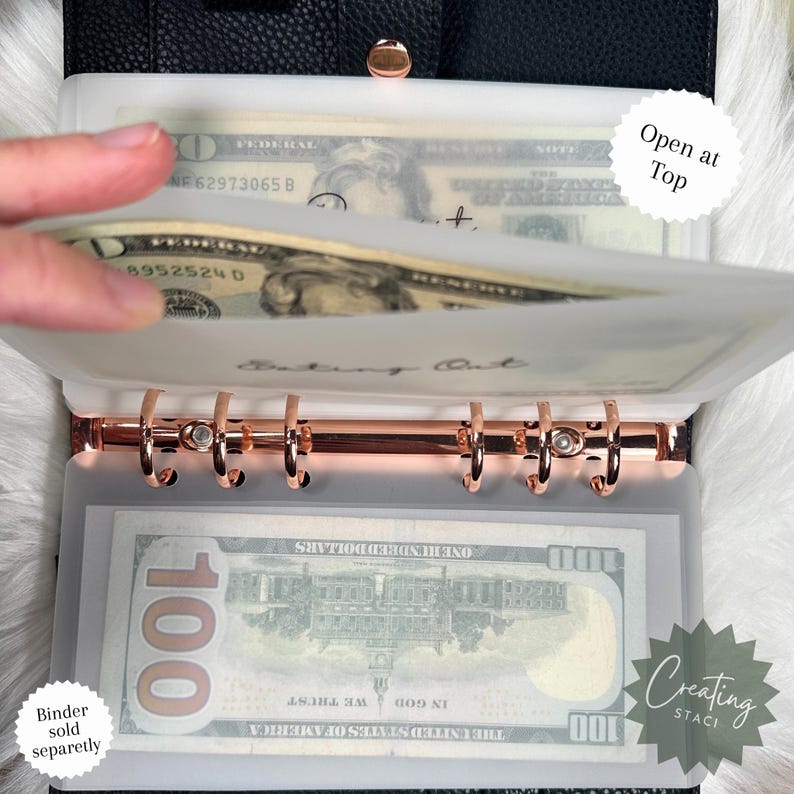 May include: A white plastic cash envelope with a rose gold binder ring system. The envelope has the words "Cash Out" written on it. The envelope is open at the top and has a $100 bill inside.