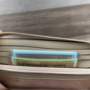 May include: A beige zip-around wallet with a gold-toned metal zipper. The interior features multiple card slots, a central zippered coin compartment, and a light-colored lining patterned with small spade symbols. A twenty-dollar bill is visible inside.