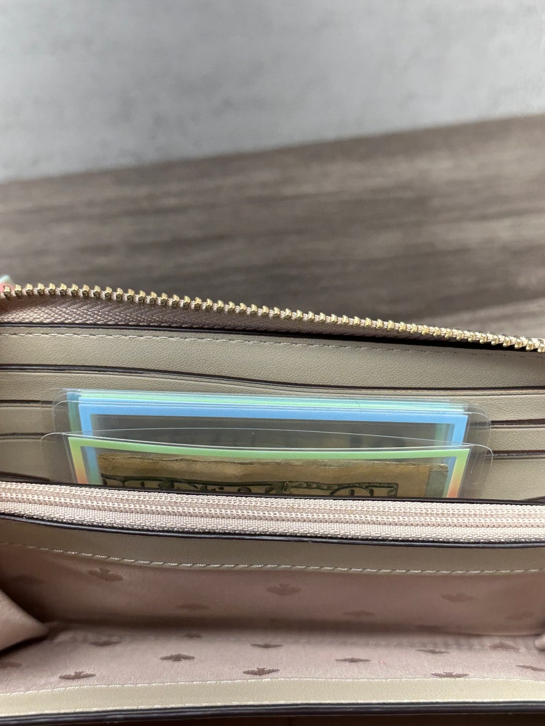 May include: A beige zip-around wallet with a gold-toned metal zipper. The interior features multiple card slots, a central zippered coin compartment, and a light-colored lining patterned with small spade symbols. A twenty-dollar bill is visible inside.