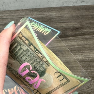 May include: A clear plastic cash envelope with iridescent edges containing a ten-dollar bill. The envelope features the word GAS printed in a pink gradient font on the front, with the word SPENDING partially visible on another envelope below.