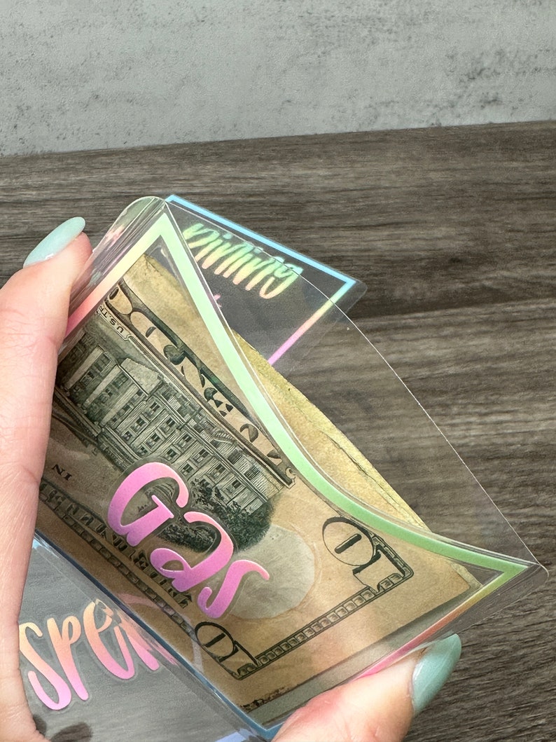 May include: A clear plastic cash envelope with iridescent edges containing a ten-dollar bill. The envelope features the word GAS printed in a pink gradient font on the front, with the word SPENDING partially visible on another envelope below.