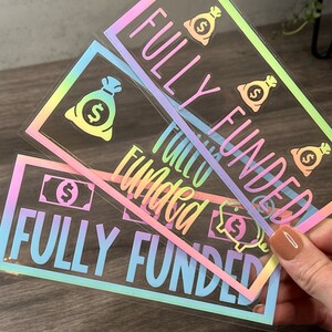 Puede incluir: Tres pegatinas rectangulares con un acabado hologr&aacute;fico de arco&iacute;ris pastel. Cada pegatina presenta el texto FULLY FUNDED en letras may&uacute;sculas negritas, acompa&ntilde;ado de iconos gr&aacute;ficos de bolsas de dinero y billetes sobre un fondo transparente.