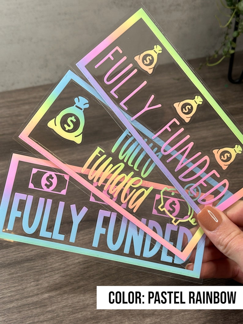 Puede incluir: Tres pegatinas rectangulares con un acabado hologr&aacute;fico de arco&iacute;ris pastel. Cada pegatina presenta el texto FULLY FUNDED en letras may&uacute;sculas negritas, acompa&ntilde;ado de iconos gr&aacute;ficos de bolsas de dinero y billetes sobre un fondo transparente.