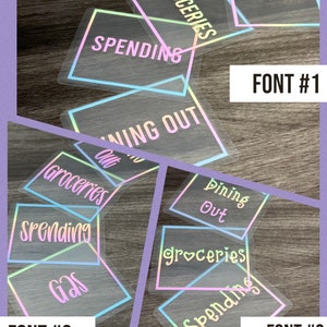 May include: A collection of rectangular transparent stickers with iridescent borders and text. The stickers feature labels including GROCERIES, GAS, SPENDING, and DINING OUT in three distinct font styles labeled FONT #1, FONT #2, and FONT #3.