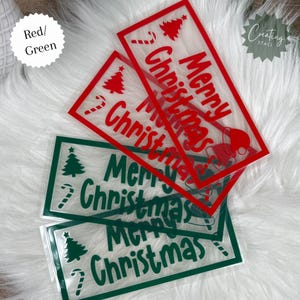 May include: Clear plastic Christmas-themed cash envelopes in red and green. The red envelopes feature a Christmas tree, candy cane, and the words "Merry Christmas." The green envelopes have similar designs. The envelopes are on a white, fluffy surface.