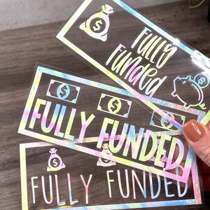 Puede incluir: Tres pegatinas rectangulares con un acabado hologr&aacute;fico de arte fluido brillante. Cada pegatina presenta el texto "FULLY FUNDED" en una fuente negrita, acompa&ntilde;ado de iconos gr&aacute;ficos de bolsas de dinero, billetes y una hucha.