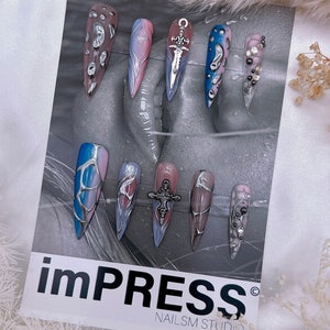 May include: A set of ten long, pointed, false nails with various designs. The nails are painted in shades of blue, pink, and white with silver accents. Some nails feature a silver sword or cross charm. The nails are displayed on a white card with the text "impRESS&reg; NAILS M STUDIO".