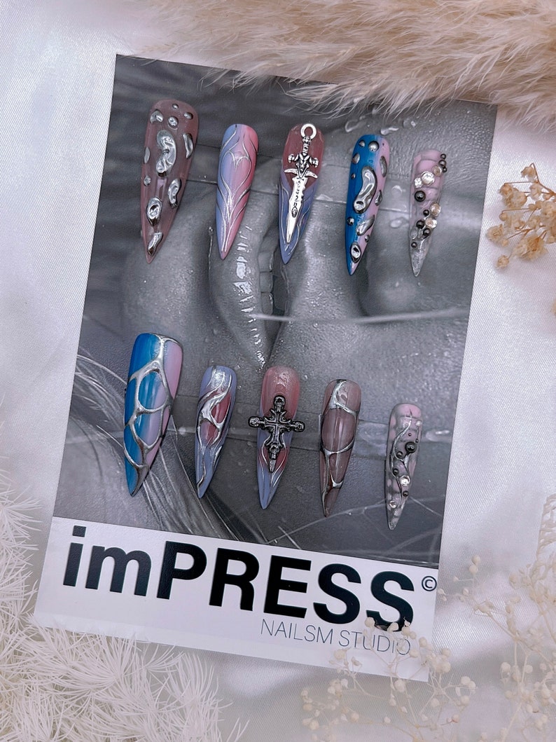 May include: A set of ten long, pointed, false nails with various designs. The nails are painted in shades of blue, pink, and white with silver accents. Some nails feature a silver sword or cross charm. The nails are displayed on a white card with the text "impRESS&reg; NAILS M STUDIO".