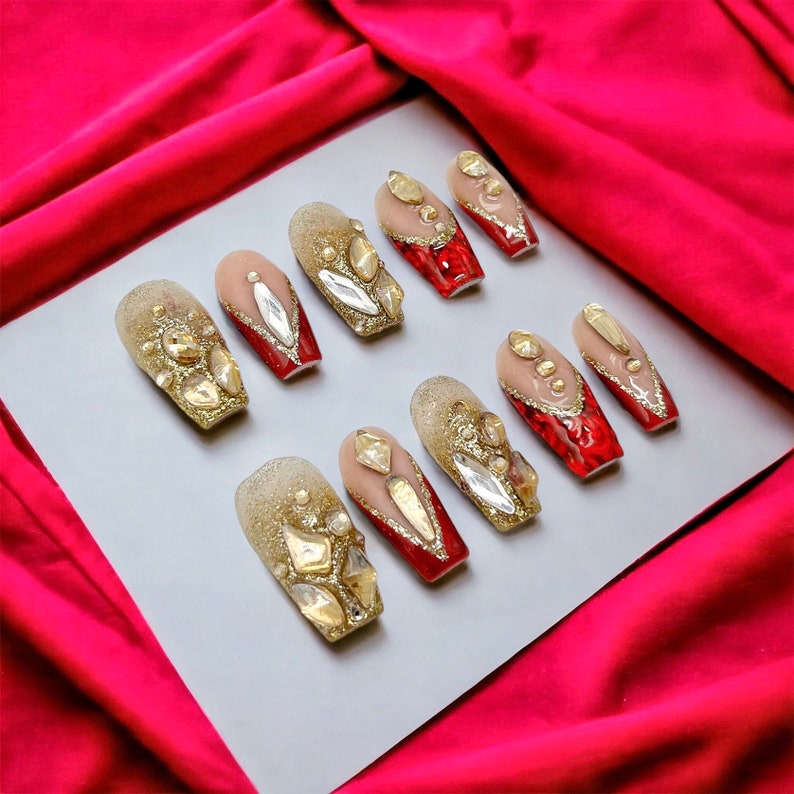 May include: Ten red and gold press-on nails with rhinestones. The nails are arranged in two rows on a white surface.