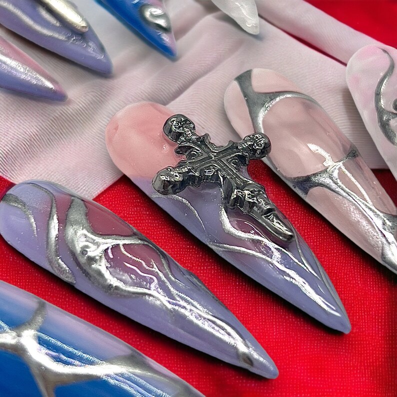 May include: Close-up of several long, pointed, false nails with a silver and purple marbled design. One nail has a silver cross charm attached to it.